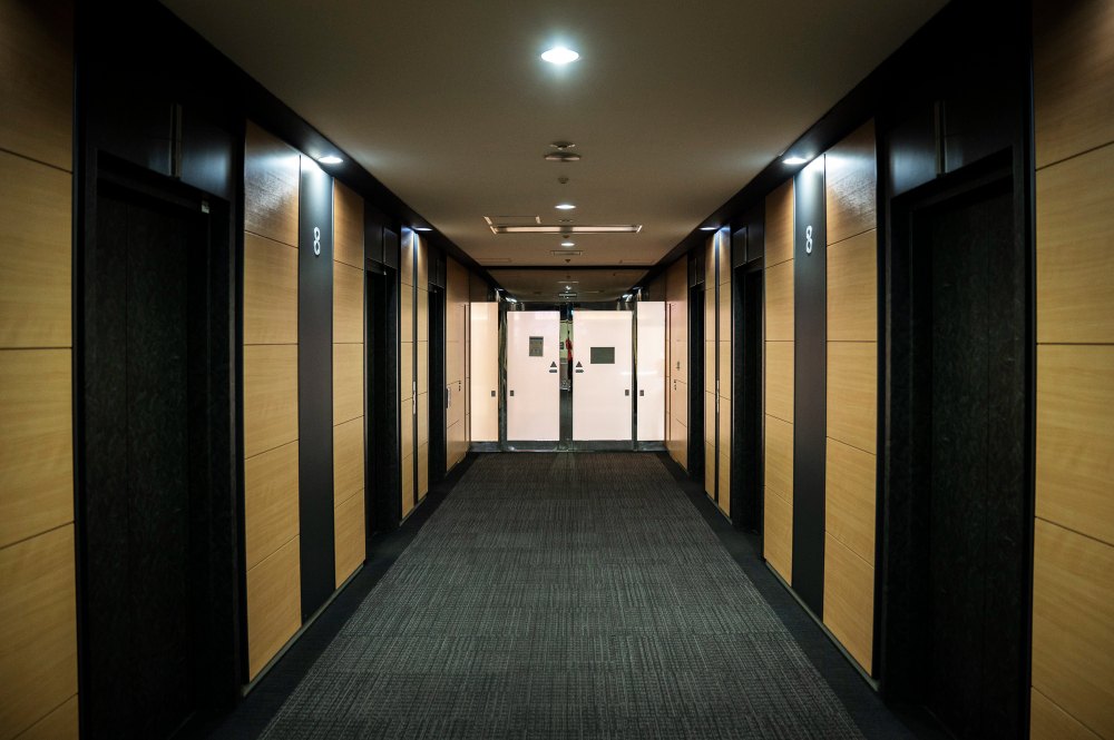 Couloir moderne d’un hôtel avec des des panneaux en bois clair sur les murs, de la moquette grise au sol, éclairé par des lumières au plafond.