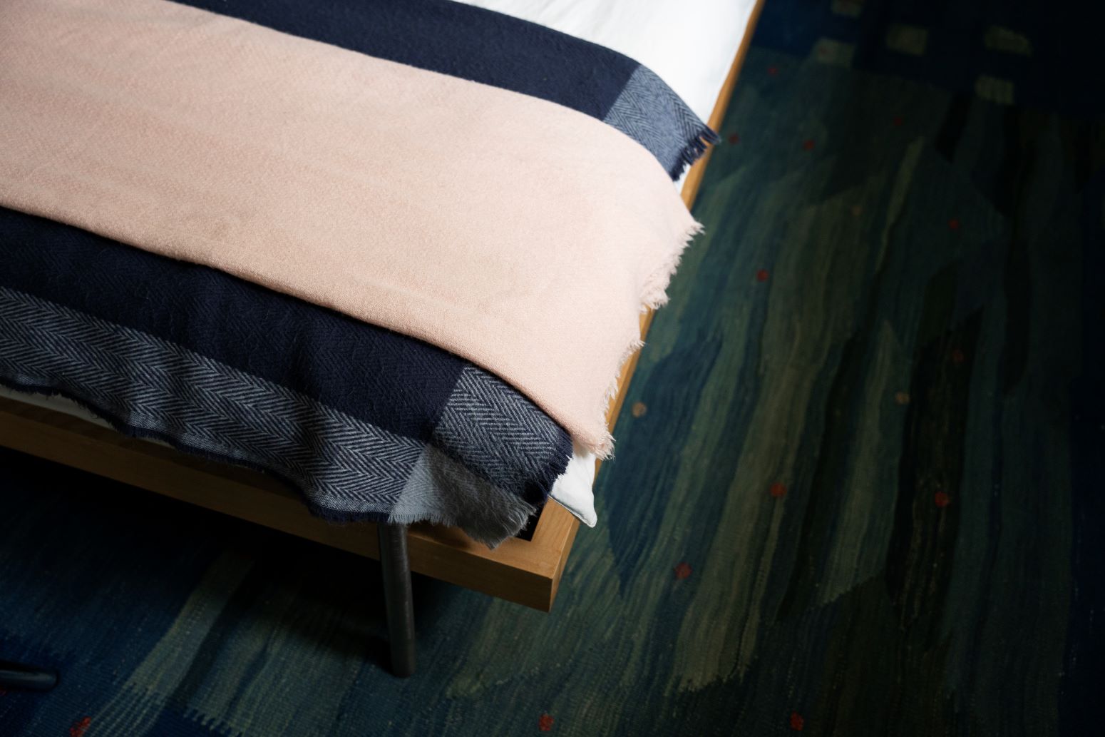 Coin de lit recouvert de plusieurs couches de tissus couvre-lits superposés. Ils sont de couleur beige rosé en haut, un tissu bleu marine au centre et un tissu à motif chevron gris et bleu en dessous.
